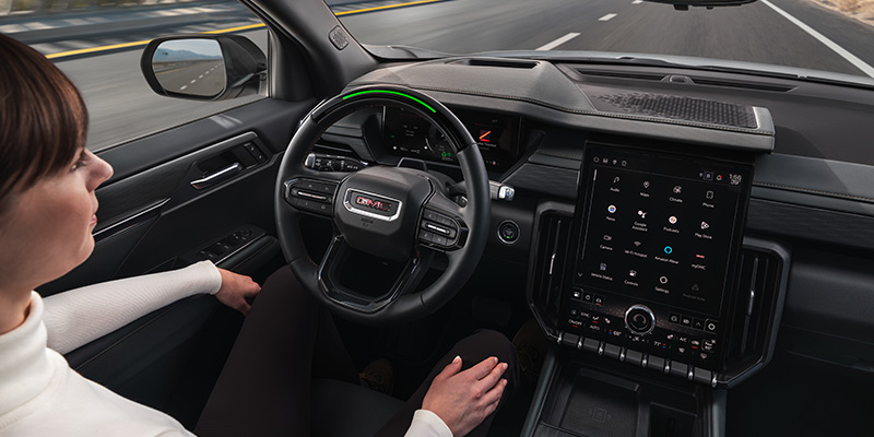 A person seated in a 2025 GMC Acadia, hands off the steering wheel, with a large display in the center console.