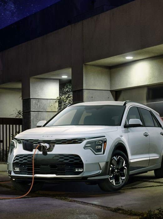 A white electric KIA SUV is parked and charging at night with bright lights above.