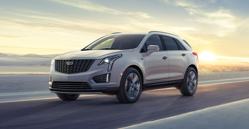 A silver 2025 Cadillac XT5 SUV driving on a road during sunset with a scenic background.