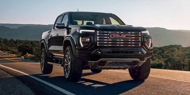 A black GMC 2025 GMC Canyon truck driving on a scenic road with mountains in the background.