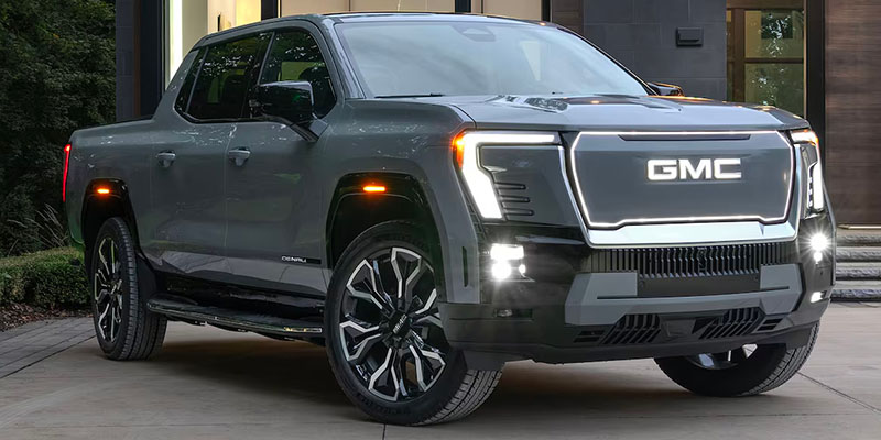 A gray 2025 GMC Sierra EV pickup truck parked on a driveway, showcasing stylish design and bright headlights.