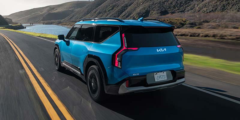 A blue 2026 Kia EV9 driving on a road next to a river, surrounded by hills.