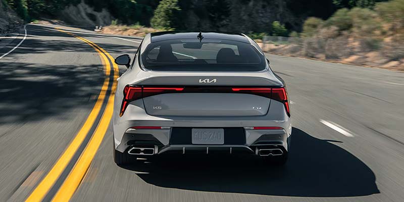 Silver Kia K5 sports sedan driving on a winding road, showcasing its sleek rear design and LED taillights.